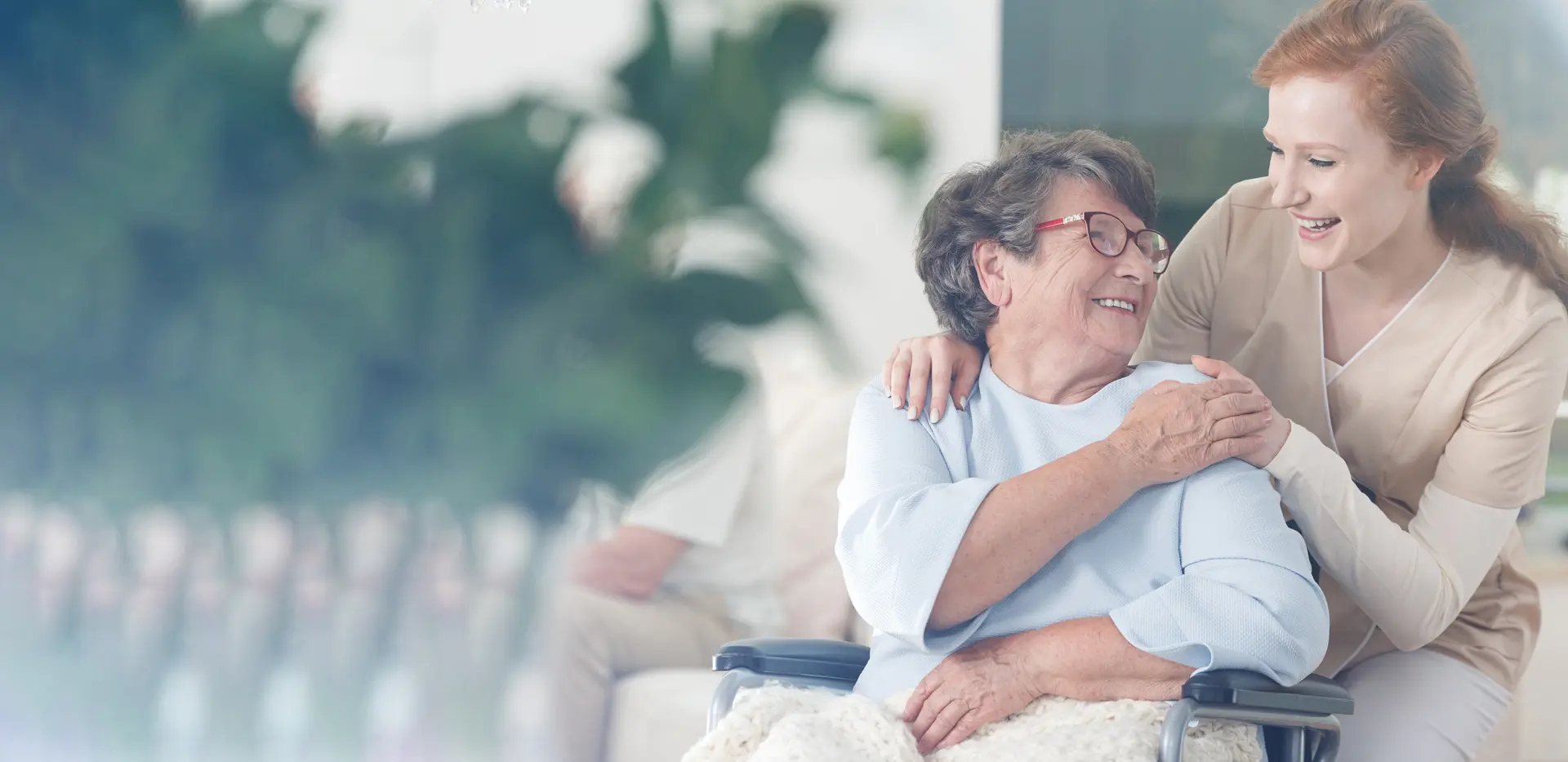 A caregiver with her arm around an older woman in a wheelchair, both smiling