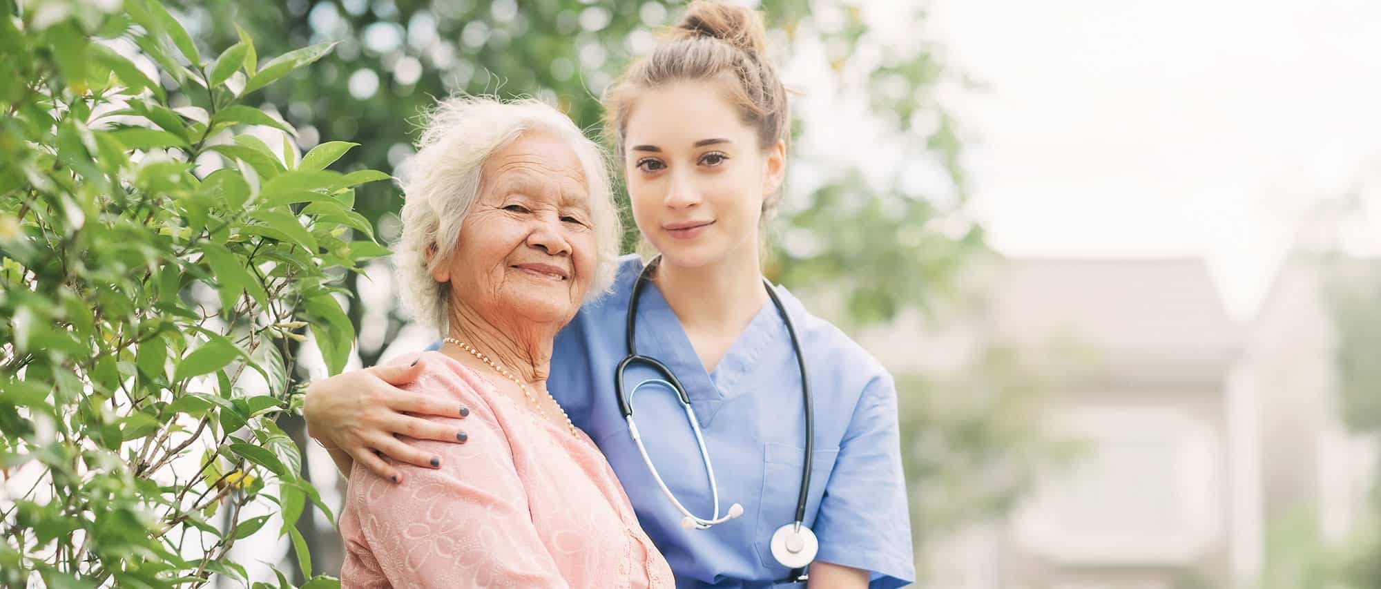 A caregiver walking outdoors with an older woman on a bright day